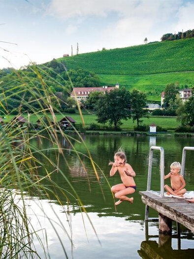 Eine Gruppe von Kindern und Erwachsenen sitzt auf einem Steg am Wasser. Im Hintergrund sind grüne Hügel und Weinberge zu sehen. | © Karin Bergmann