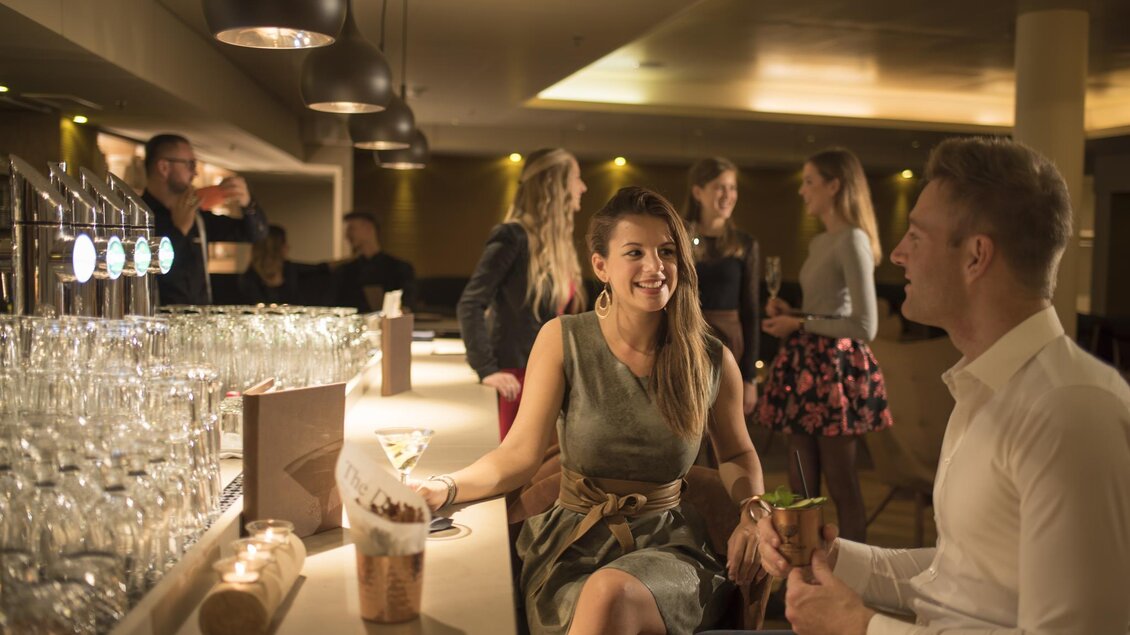 <p>Eine elegante Bar mit fröhlichen Menschen. Eine Frau unterhält sich lächelnd mit einem Mann, während andere Gäste im Hintergrund stehen. Sunny Bar in Bad Loipersdorf.</p> | © Thermenhotel Das Sonnreich