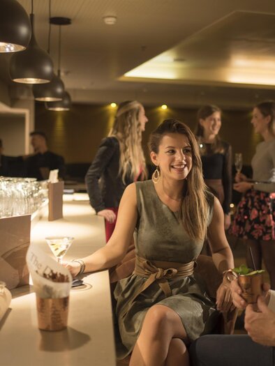 <p>Eine elegante Bar mit fröhlichen Menschen. Eine Frau unterhält sich lächelnd mit einem Mann, während andere Gäste im Hintergrund stehen. Sunny Bar in Bad Loipersdorf.</p> | © Thermenhotel Das Sonnreich