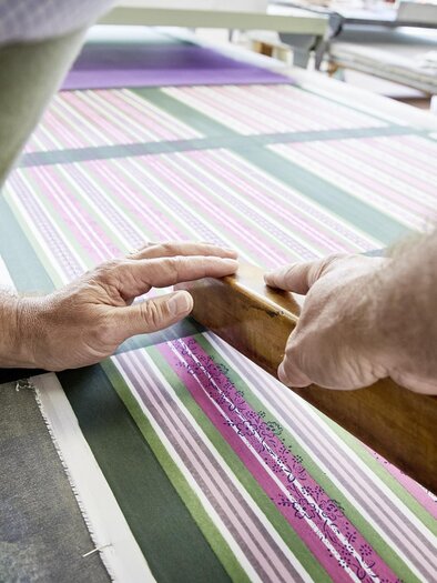 A craftsman is working on a striped fabric sample with a wooden tool. The hands are placed on the table as they work precisely. | © Handdrucke Sekyra-Helge Kirchberger