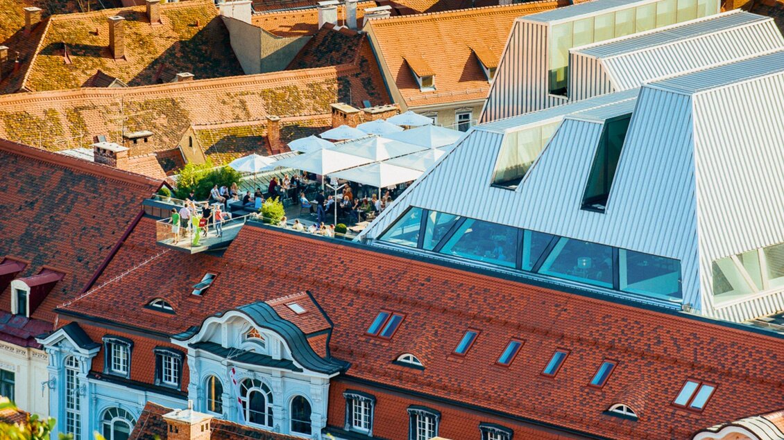 Ein Blick auf moderne und historische Architektur mit roten Dächern. Auf der oberen Etage befinden sich Sonnenschirme und eine Terrasse, die für Gäste eingerichtet ist. | © Tagescafé Freiblick