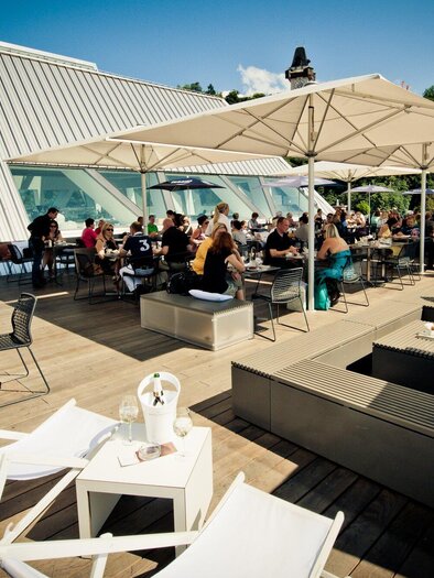 Eine belebte Dachterrasse mit vielen Gästen, die an Tischen sitzen. Schirme spenden Schatten, und die Atmosphäre ist gemütlich und einladend. | © K&Ö - Lupispuma