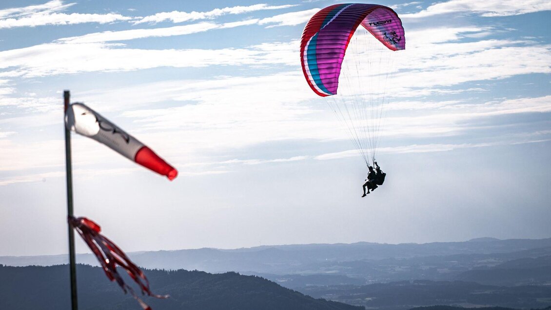<p>Ein Paragleiter schwebt durch den Himmel über dem Salzkammergut mit einem bunten Schirm. Im Vordergrund weht eine Fahne im Wind.</p> | © Flugschule Salzkammergut