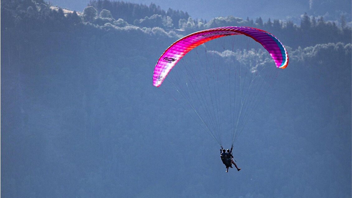 <p>Ein Paraglider schwebt elegant über dem Ausseerland. Im Hintergrund sind grüne Hügel mit Bäumen zu sehen.</p> | © Flugschule Salzkammergut