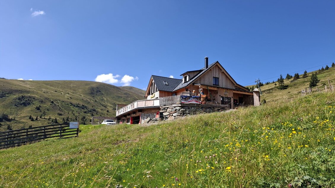 Ein gemütliches Haus auf einer Wiese, umgeben von grünen Hügeln. Der Himmel ist klar und blau. | © Tanzstatt Hütte
