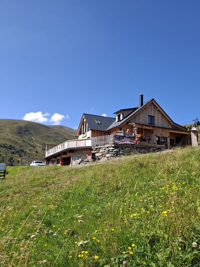 Ein gemütliches Haus auf einer Wiese, umgeben von grünen Hügeln. Der Himmel ist klar und blau. | © Tanzstatt Hütte