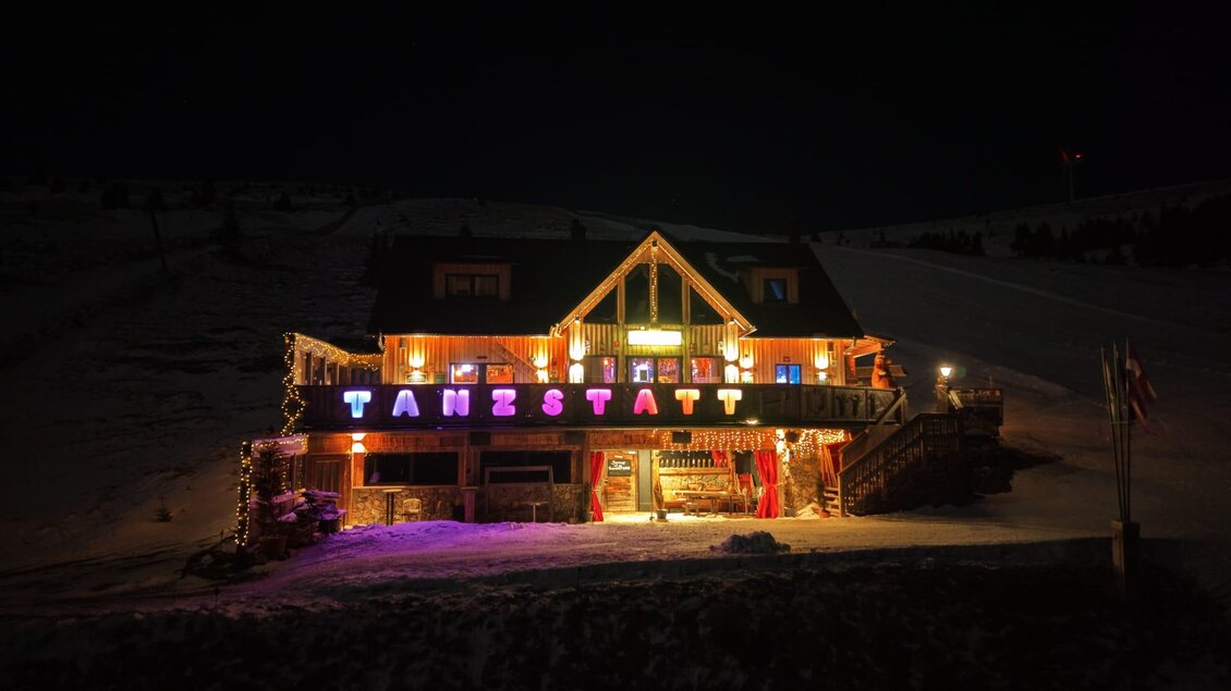 Eine beleuchtete Berghütte bei Nacht, umgeben von Schnee. Der Schriftzug "TANZSTATT" ist farbenfroh und gut sichtbar. | © Tanzstatt Hütte