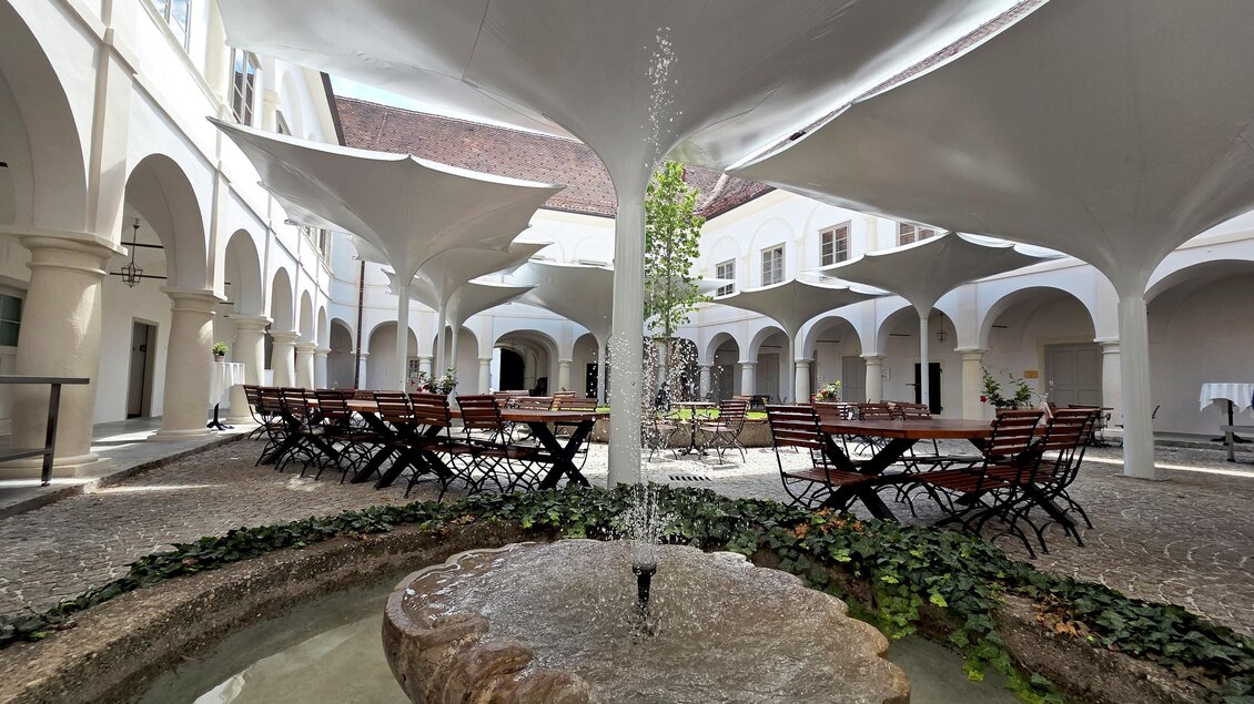 Ein schöner Innenhof mit Tischen und Stühlen aus Holz. In der Mitte sprudelt ein Brunnen, umgeben von Pflanzen. | © Schloss Retzhof
