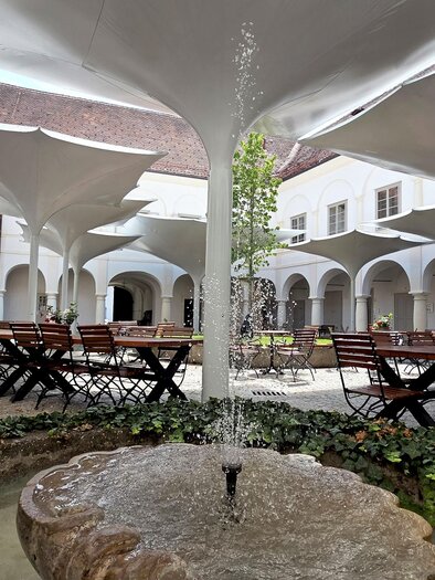 Ein schöner Innenhof mit Tischen und Stühlen aus Holz. In der Mitte sprudelt ein Brunnen, umgeben von Pflanzen. | © Schloss Retzhof