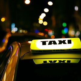Ein Taxi mit leuchtendem Schild auf einer nächtlichen Straße. Die Umgebung ist dunkel, aber mit farbigen Lichtern beleuchtet. | © Unsplash maik-winnecke