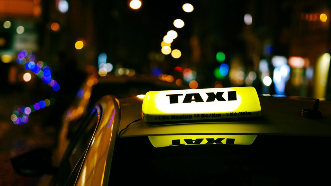 Ein Taxi mit leuchtendem Schild auf einer nächtlichen Straße. Die Umgebung ist dunkel, aber mit farbigen Lichtern beleuchtet. | © Unsplash maik-winnecke