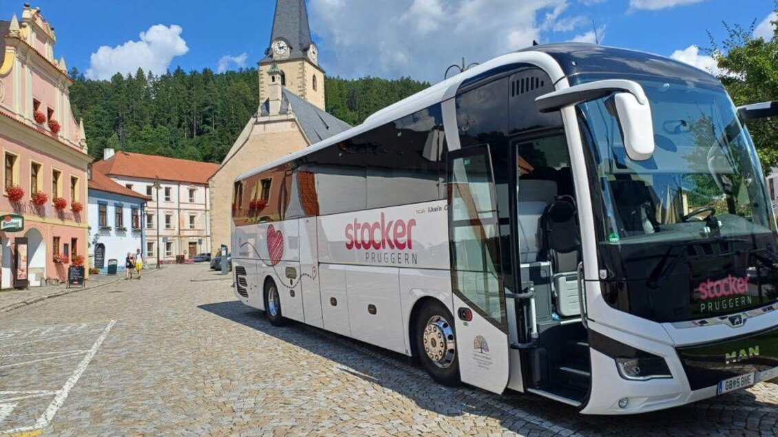 Ein moderner Reisebus steht auf einem gepflasterten Platz in einer malerischen Stadt. Im Hintergrund sind historische Gebäude und ein Kirchturm zu sehen. | © Stocker Reisen