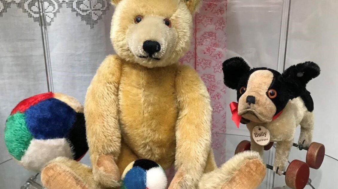 Ein gelbes Plüschbärchen sitzt mit bunten Bällen daneben. Neben ihm steht ein kleiner Plüschhund auf Rädern. | © Teddy und Spielzeugmuseum Lannach
