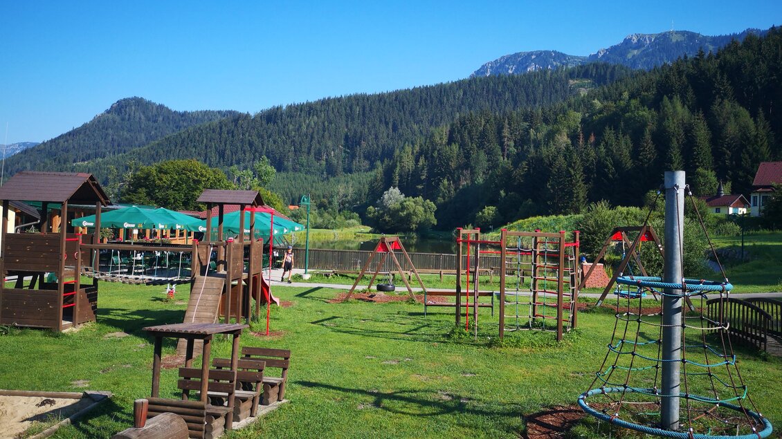 Ein Spielplatz mit verschiedenen Spielgeräten steht vor einer grünen Wiese und sanften Hügeln. Im Hintergrund sind Bäume und Berge zu sehen, unter einem klaren blauen Himmel.