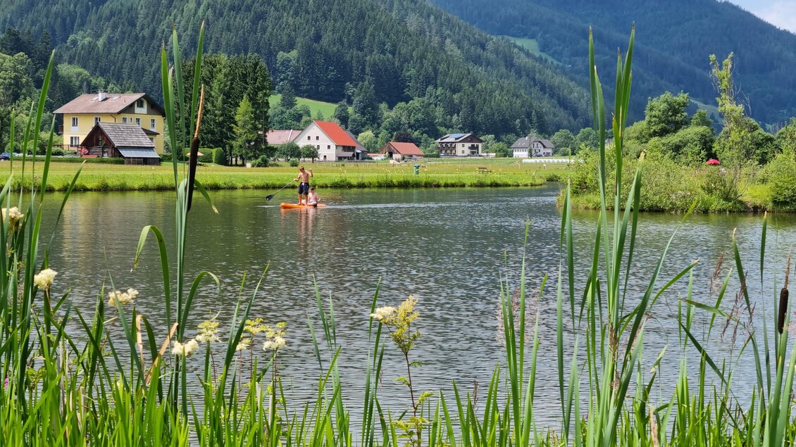 Ein ruhiger See umgeben von Wiesen und sanften Hügeln. Im Vordergrund sind hohe Gräser und im Hintergrund paddelt jemand auf dem Wasser. | © Naturpark Mürzer Oberland, Andreas Steininger
