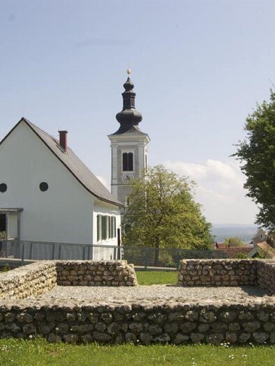 A historical building with a bell tower stands next to a stone ruin. The surroundings are green with several trees and a clear sky. | © Maria Schoiswohl