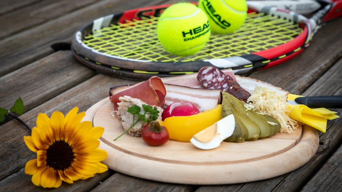 Eine Holzplatte mit verschiedenen Wurst- und Käsesorten, kleinen Gurken und einer Blume. Im Hintergrund liegen Tennisbälle und ein Schläger. | © TV Region Graz-Lunghammer