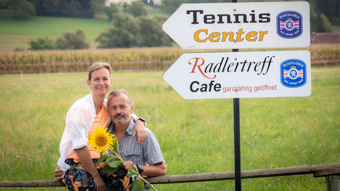 Ein Paar posiert fröhlich vor einem Wegweiser, der auf ein Tenniscenter und ein Radleitreff-Café hinweist. Im Hintergrund ist eine grüne Landschaft mit sanften Hügeln zu sehen. | © TV Region Graz - Lunghammer