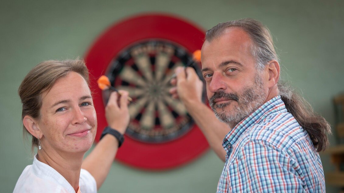 Ein Paar steht vor einer Dartscheibe und lächelt. Beide zeigen mit Darts auf das Ziel. | © TV Region Graz - Lunghammer
