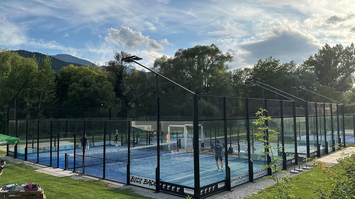 Ein Padelplatz umgeben von einem grünen Rasen und Bäumen. Im Hintergrund sind Spieler aktiv am Spielen, und der Himmel ist teils bewölkt. | © Anna-Lena König