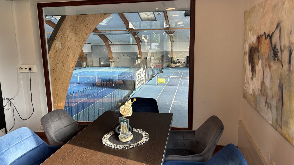 Ein gemütlicher Raum mit einem Tisch und blauen Stühlen. Durch das Fenster ist ein Tennisplatz sichtbar. | © Anna-Lena König