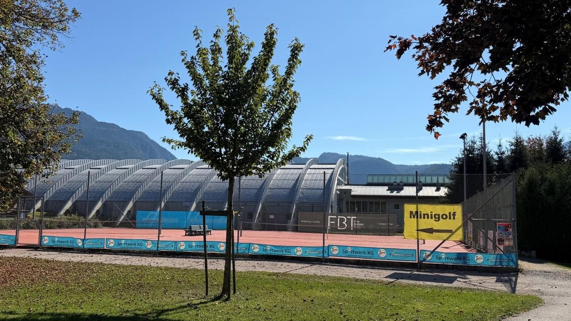 Eine Tennisanlage mit einem Minigolfplatz im Vordergrund und einem klaren blauen Himmel. Im Hintergrund sind Berge und eine grüne Landschaft zu sehen. | © Tennishalle Bad Mitterndorf
