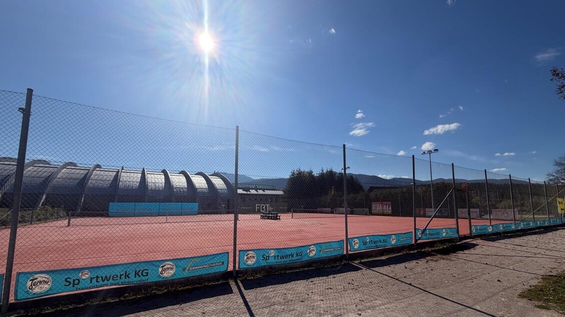 Ein Tennisplatz mit rotem Belag und einem klaren blauen Himmel. Die Umgebung ist gut beleuchtet und freundlich. | © Tennishalle Bad Mitterndorf