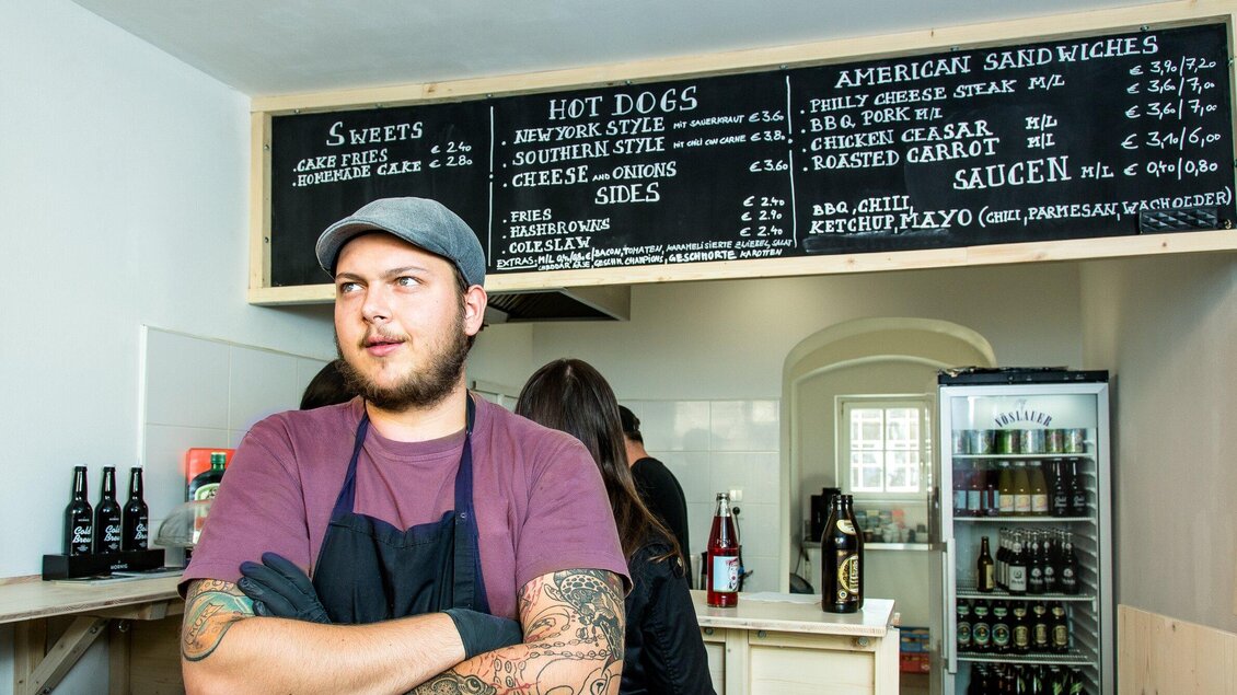 Ein junger Mann steht in einem Restaurant mit einem Menü über ihm. Das Menü enthält verschiedene Snacks und Gerichte wie Hot Dogs und Sandwiches. | © The Hungry Heart