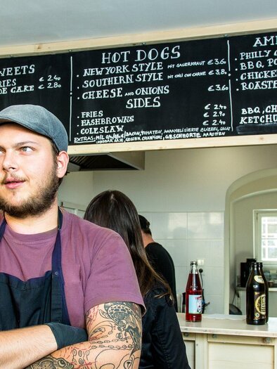 Ein junger Mann steht in einem Restaurant mit einem Menü über ihm. Das Menü enthält verschiedene Snacks und Gerichte wie Hot Dogs und Sandwiches. | © The Hungry Heart