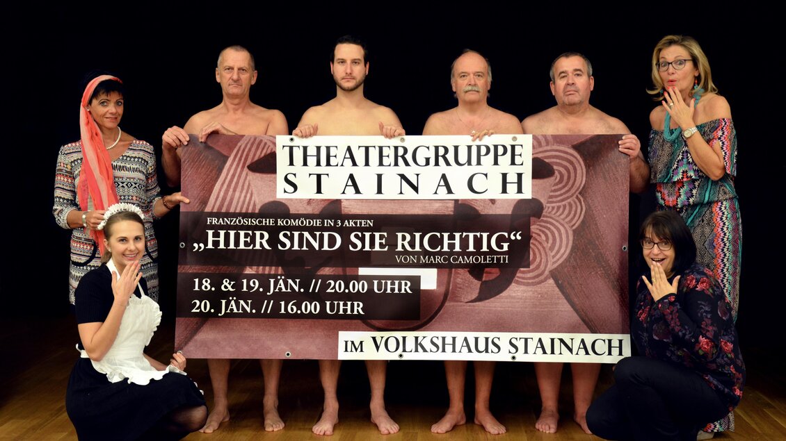 Eine Theatergruppe präsentiert ein Plakat für eine Komödie. Die Darsteller posieren kreativ und humorvoll in einem Theaterraum.
