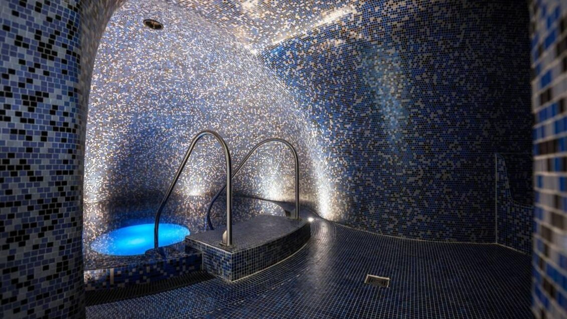 Eine entspannende Wellness-Oase mit glitzernden blauen Fliesen. Im Hintergrund ist ein kleiner Pool mit sanftem Licht zu sehen. | © Hotel & Therme NOVA