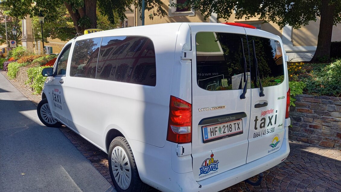 Ein weißes Taxi-Fahrzeug steht an einer Straßenkante. Der Hintergrund zeigt grüne Bäume und eine ruhige Wohnstraße. | © Thermen- & Vulkanland