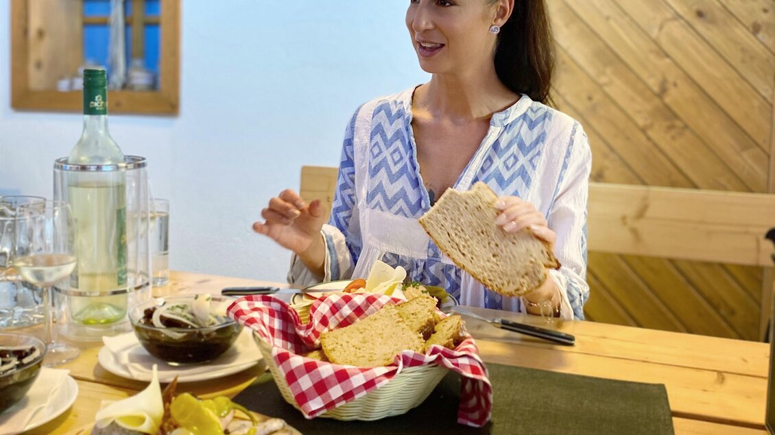 Eine lächelnde Frau sitzt an einem Holztisch, umgeben von verschiedenen Speisen. Auf dem Tisch sind Brot, Wurst und frisches Gemüse angerichtet. | © silviaschreibt.de
