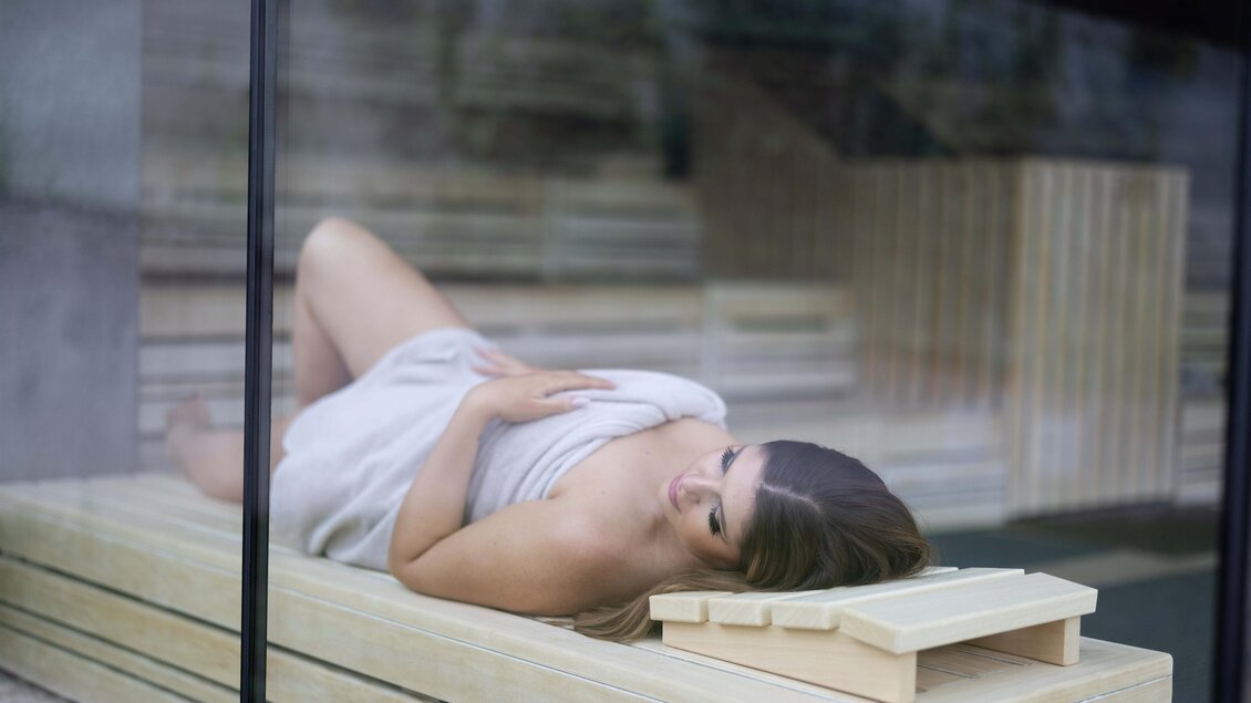 <p>Eine Person liegt entspannt auf einer Holzliege in einem modernen Raum. Der Raum hat große Fenster und eine minimalistische Gestaltung. Thermenresort Loipersdorf in Bad Loipersdorf.</p> | © TRL