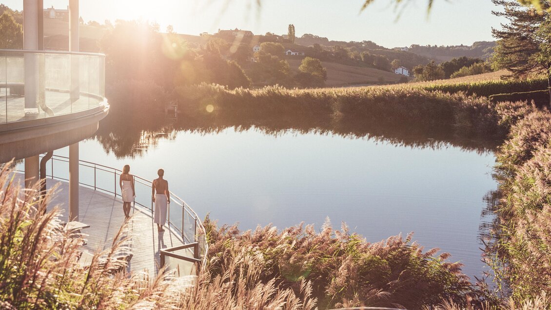 <p>Eine ruhige Seenlandschaft bei Sonnenuntergang. Zwei Personen spazieren entlang der Uferpromenade. Thermenresort Loipersdorf in Bad Loipersdorf.</p> | © TRL