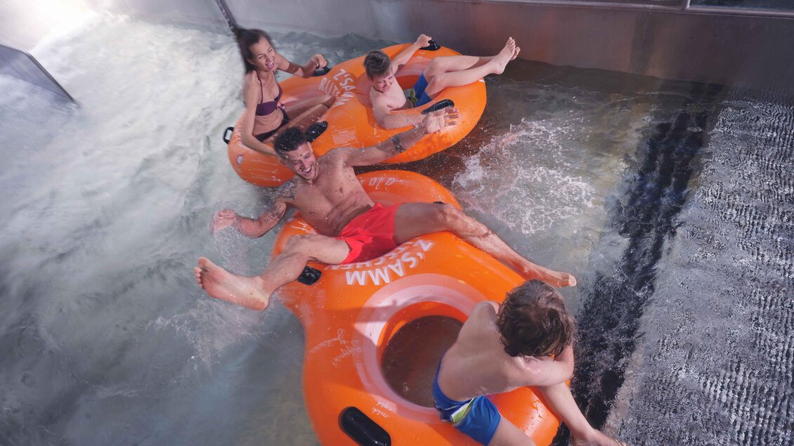 <p>Vier Kinder fahren in orangefarbenen Schwimmreifen auf einer Wasserrutsche. Sie haben Spaß und spritzen Wasser um sich herum. Thermenresort Loipersdorf in Bad Loipersdorf.</p> | © TRL