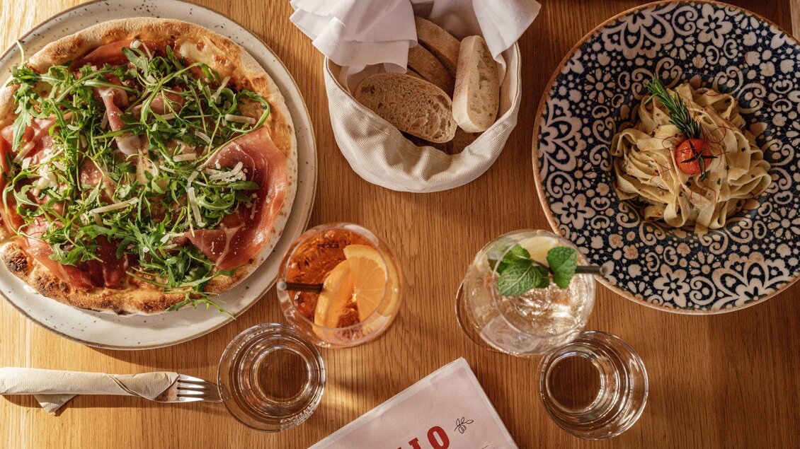 <p>Eine köstliche Pizza mit Rucola und Schinken neben einem Teller Pasta. Dazu gibt es frisches Brot und Getränke auf einem Holztisch. Thermenresort Loipersdorf in Bad Loipersdorf.</p> | © TRL
