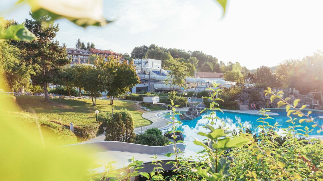 <p>Eine grüne Gartenlandschaft mit einem blauen Pool und zahlreichen Bäumen. Im Hintergrund sind Gebäude und sanfte Hügel sichtbar. Thermenresort Loipersdorf in Bad Loipersdorf.</p> | © TRL