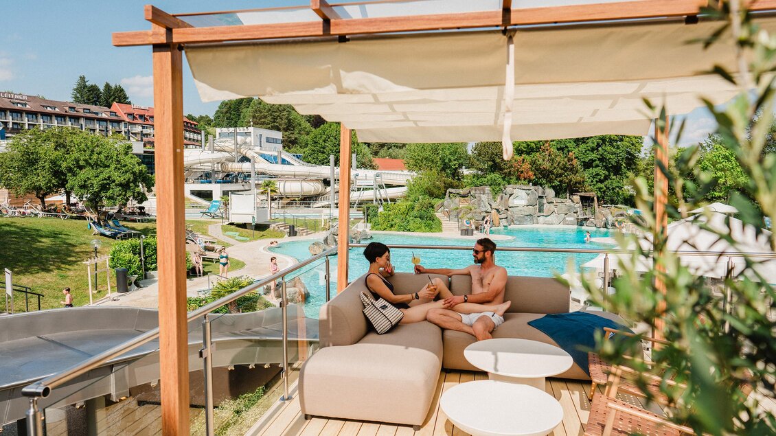 <p>Eine gemütliche Terrasse mit einer Lounge-Sitzgruppe und zwei Personen, die sich unterhalten. Im Hintergrund sind ein Schwimmbad und Rutschen zu sehen. Thermenresort Loipersdorf in Bad Loipersdorf.</p> | © 5komma5sinne/motionAds