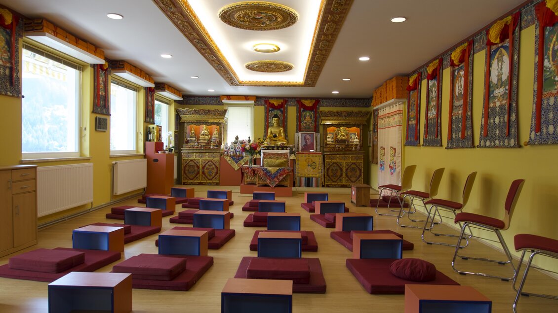 Ein meditativ gestalteter Raum mit roten Sitzkissen und bunten Wandbehängen. In der Mitte befindet sich ein Altar mit buddhistischen Statuen und Blumen. | © Tibetzentrum Institut