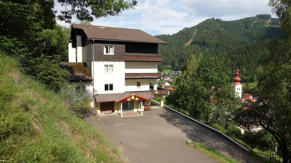 Ein modernes Gebäude in einer bergigen Umgebung. Im Hintergrund sind grüne Wälder und ein Kirchturm sichtbar. | © Tibetzentrum Institut