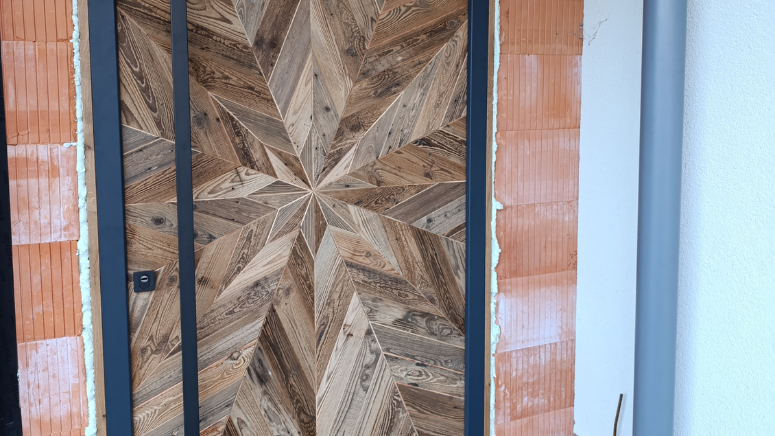Eine kunstvoll gestaltete Holztür mit einem symmetrischen Muster aus verschiedenen Holzstücken. Die Tür ist in einem modernen Gebäudeeingang eingebaut. | © Tischlerei Egger