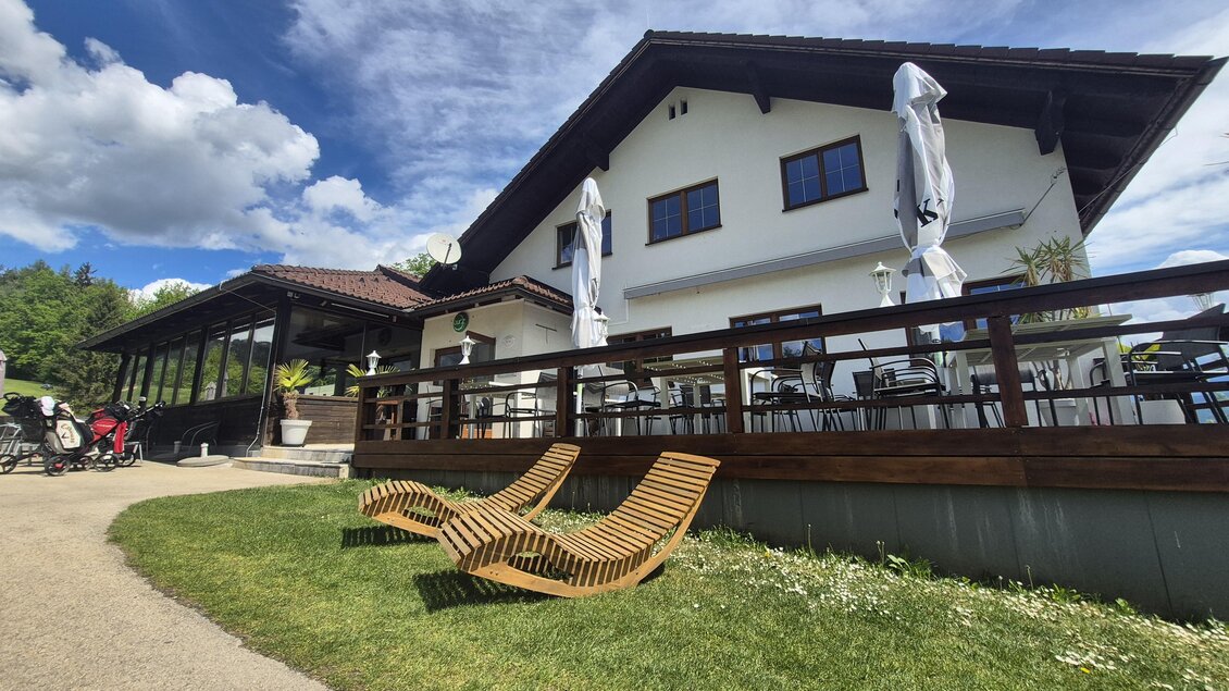 Ein schönes Gebäude mit einer Terrasse und Sonnenschirmen. Im Vordergrund stehen zwei Holzliegestühle auf einer grünen Wiese. | © Andrea Stelzer