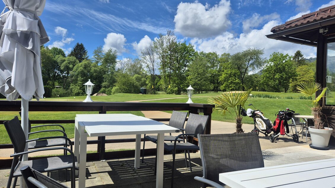 Eine gemütliche Terrasse mit einem weißen Tisch und Stühlen. Im Hintergrund sieht man Golfplätze und grüne Bäume unter einem blauen Himmel. | © Andrea Stelzer
