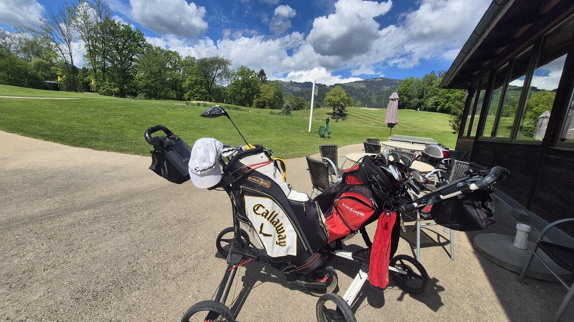 Zwei Golfcaddies stehen auf einem Parkplatz in der Nähe eines Golfplatzes. Der Himmel ist blau mit einigen Wolken und im Hintergrund sind Bäume zu sehen. | © Andrea Stelzer