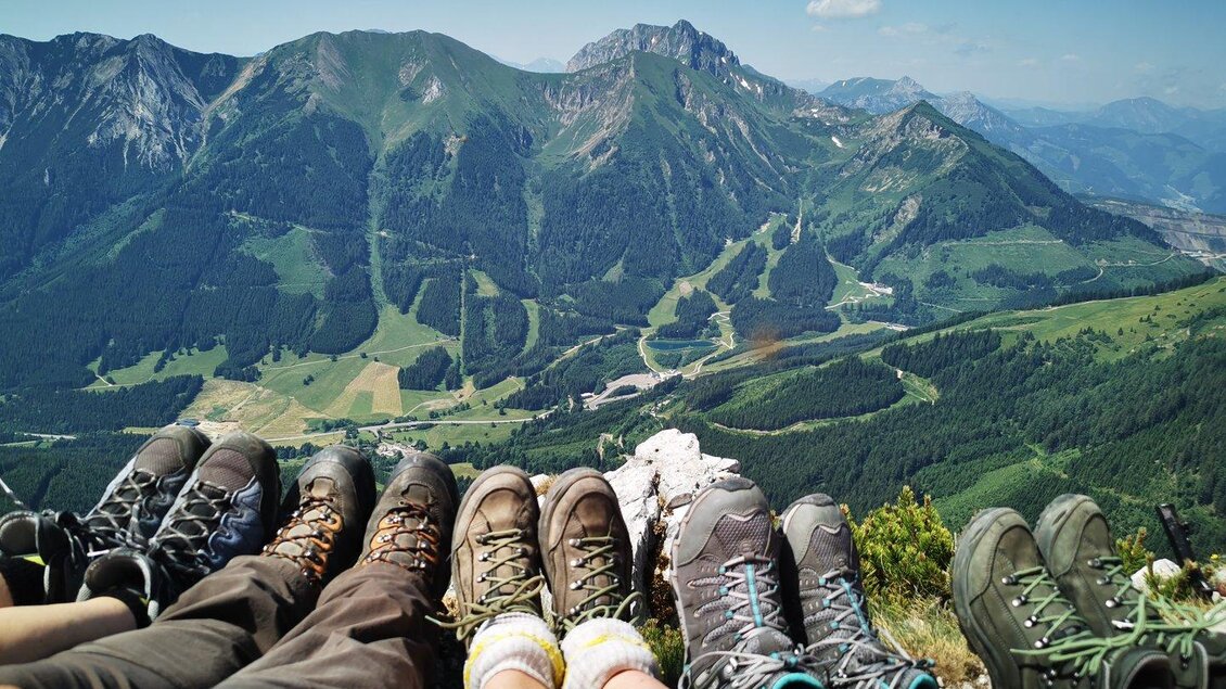 Eine Gruppe von Menschen sitzt auf einem Berggipfel und zeigt ihre Wanderschuhe. Im Hintergrund erstreckt sich eine beeindruckende Berglandschaft mit grünen Wiesen und blauen Himmel. | © TVB Erzberg-Leoben