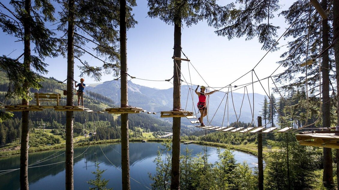 Ein Hochseilgarten hoch in den Bäumen. Im Hintergrund ist ein klarer See und eine wunderschöne Berglandschaft zu sehen. | © alpfox.com