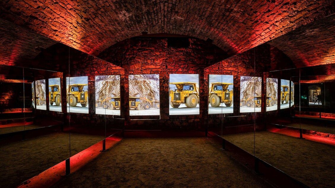 Ein modernes Kunstinstallationsraum mit roten Lichtern und großen Bildschirmen. Die Bildschirme zeigen Baustellenfahrzeuge in einer Umgebung mit Sand. | © Museumsverbund Betriebsges.m.b.H.