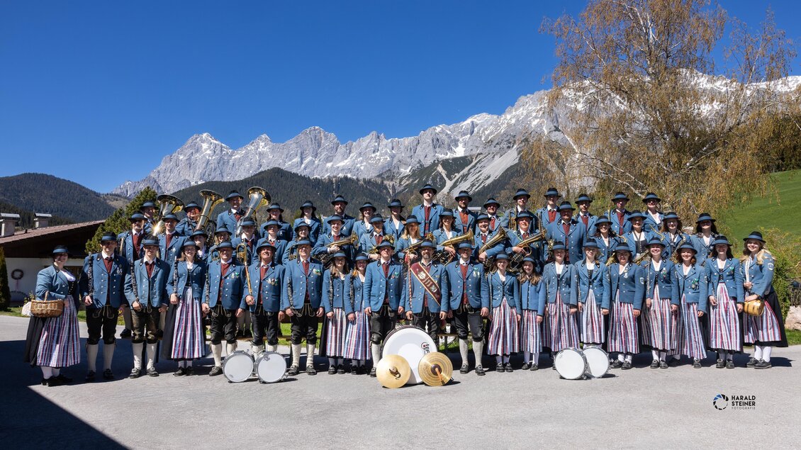 Eine große Gruppe von Menschen steht in traditionellen Trachten vor einer beeindruckenden Bergkulisse. Sie halten Musikinstrumente und lächeln in die Kamera. | © Harald Steiner