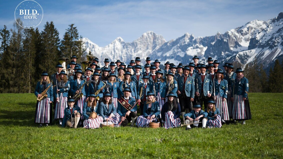Eine große Gruppe von Menschen steht auf einem grünen Feld vor schneebedeckten Bergen. Sie tragen traditionelle Trachten und halten Musikinstrumente. | © Bildwerk Fotografie